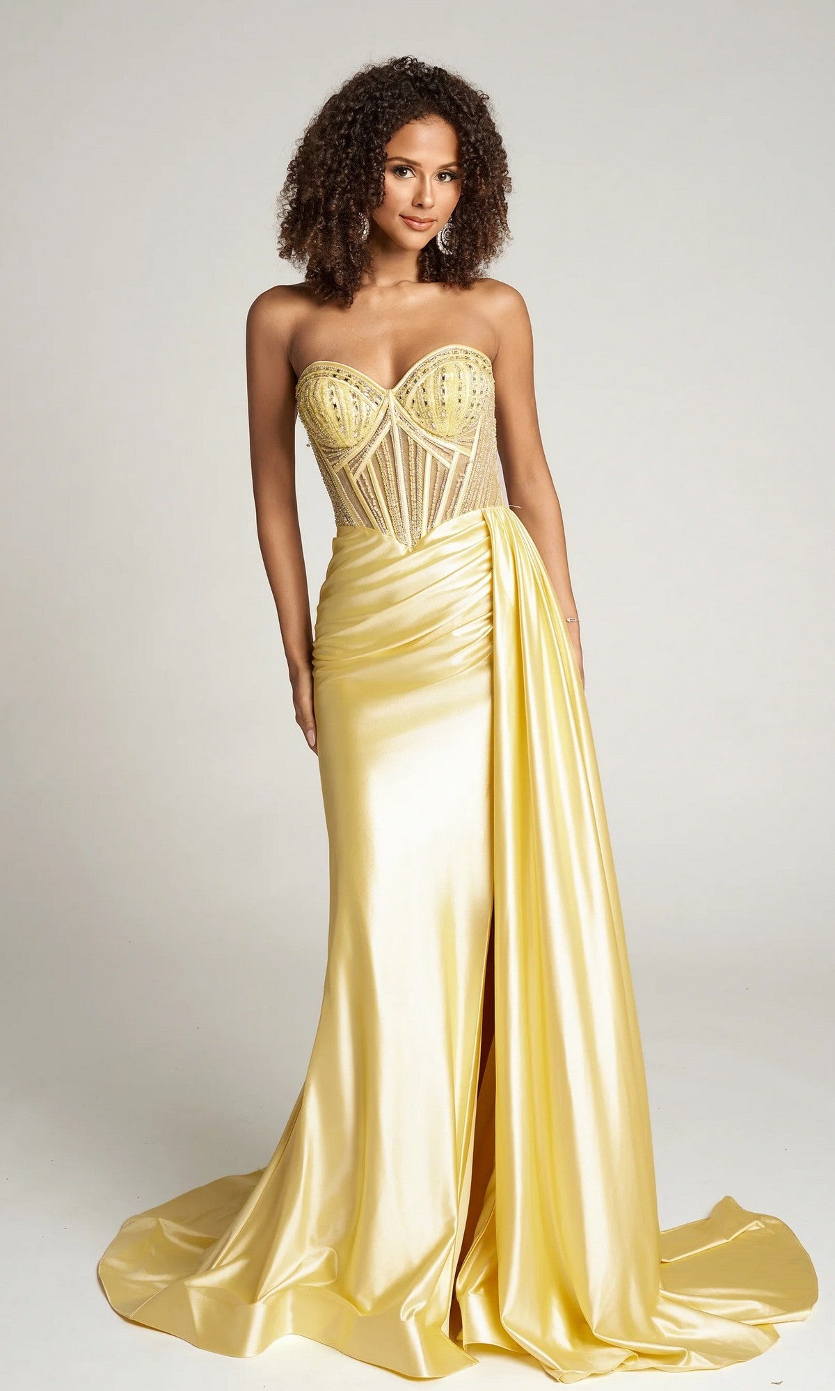 A woman wears the Portia and Scarlett PS26056 long formal dress in yellow, featuring a strapless design with a sheer corset bodice and a draped, ruched skirt highlighted by a front slit.